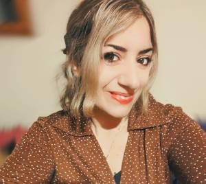 A woman with medium-length blonde hair styled in waves, wearing a brown polka dot blouse, smiling at the camera.